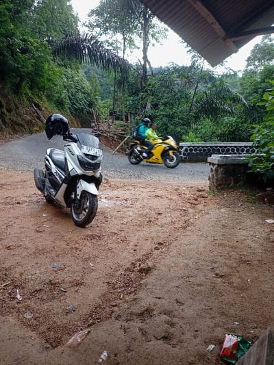 The image shows a white Yamaha motorcycle parked on a dirt road in Cisewu, Indonesia (latitude: -7.411, longitude: 107.542). The motorcycle is facing away from the camera. A yellow motorcycle with two people on it is driving away from the camera in the distance on the same dirt road. The road appears to be unpaved and wet. There are trees and bushes on both sides of the road. The image was taken from a low angle, possibly from the ground or from a low platform. There is a roof overhead in the background. The camera is pointing toward the yellow motorcycle in the distance. The image is taken in a rural setting, and the focus is on the motorcycles.