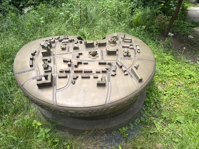 The image shows a detailed architectural model of the city of Kolomyia, Ukraine, situated outdoors in a grassy area. The model, made of a metallic material, depicts various buildings and street layouts, arranged on a circular, slightly elevated platform. The platform itself has text inscribed on its curved side, partially obscured by grass. The surrounding environment is natural, with abundant green grass, various plants, and a tree trunk visible on the right side of the frame. The lighting suggests it is daytime with some sunlight filtering through. There are no people visible in the image.
