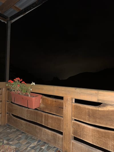 The image depicts a nighttime view from a wooden balcony or porch in Volkonka, Russia. In the foreground, a brown planter box holds red and white flowers. The balcony is enclosed by a wooden railing constructed from horizontal planks. Below the railing, a patterned tiled floor is visible. The background is dominated by the dark silhouette of mountains against a dark sky. The lighting suggests it is night, with faint light sources possibly visible in the distance. The scene appears to be of a residential or guest house balcony overlooking a natural landscape.