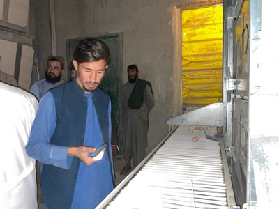 The image depicts an indoor scene within an egg production facility in Shuyene Vosta, Afghanistan. In the foreground, a young man with dark hair and a short beard, dressed in a blue long-sleeved shirt and a dark blue waistcoat, is intently observing a smartphone held in his right hand. To his right, a white-ribbed conveyor belt is visible, carrying several light brown eggs. This conveyor belt extends from a larger metallic industrial machine positioned on the right side of the frame. In the background, several other men are present, observing the activity. One man with a dark beard and a turban is partially visible to the left, and another man with a dark beard in a dark traditional garment stands further back on the right. An opening in the rear wall, possibly a doorway, is covered by a bright yellow, folded tarpaulin or curtain, which appears to be a source of ambient light. The walls of the facility are constructed of rough concrete or plaster, suggesting a utilitarian environment.