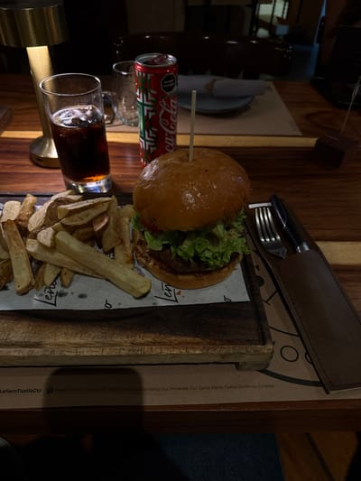 An overhead shot presents a burger and fries meal on a wooden serving board. A Coca-Cola can rests in a glass holder next to a glass of cola. Cutlery in a leather holder sits above the meal. The board's paper liner is branded with "Leñero". The board's edge is printed with "LeñeroTuxtlaGtz" and an address in Tuxtla Gutiérrez, Chiapas. The scene is warmly lit, casting shadows and highlighting the wood grain of the table surface.