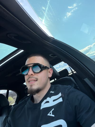 A medium shot of a light-skinned man with a short haircut and beard is taken inside a car looking upward through a sunroof. He is wearing sunglasses and a black t-shirt with large white lettering. The sunroof reveals a blue sky with sparse clouds. The car interior is black. The location cannot be determined to be Biaga, Bulgaria, from the image.
