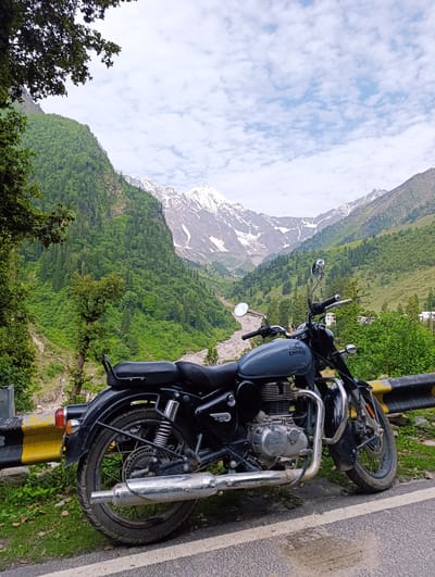 A grey Royal Enfield Classic 350 motorcycle is parked on the side of a paved road in Dhundi, India. The motorcycle is positioned at an angle, with its front wheel slightly turned towards the left. It is facing away from the viewer, with the rear of the bike closer to the camera. A yellow and black striped barrier runs along the edge of the road, behind the motorcycle.

The background is dominated by a lush green valley, with steep, tree-covered mountains on either side. In the distance, snow-capped peaks rise against a partly cloudy sky. The weather appears overcast, but the clouds are scattered, allowing some sunlight to illuminate the landscape. A riverbed is visible in the valley, with rocks and a small stream. The vegetation is dense, with a mix of trees and shrubs. The overall impression is one of a scenic, mountainous location, likely a popular spot for motorcycle riders. There are no people visible in the image.