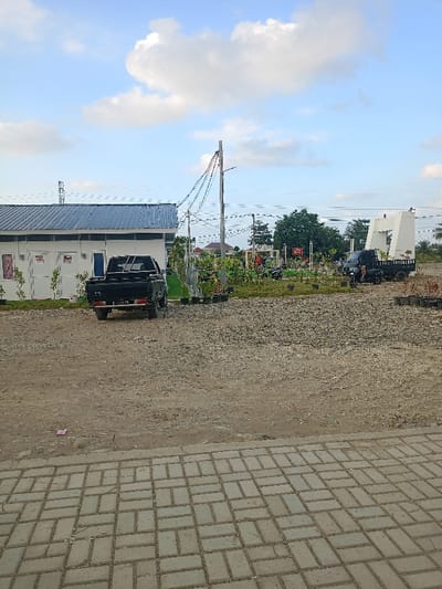 The image displays an outdoor scene in Meureudu, Indonesia, under a blue sky with scattered white cumulus clouds. The immediate foreground features a light grey paver block surface, arranged in a basket-weave pattern. Adjacent to this, the ground is unpaved, composed of dirt and gravel, with a small piece of pink litter visible on the left.

In the midground, a single-story white building with a dark blue corrugated metal roof is present on the left. It has multiple white doors and small rectangular windows. A black pickup truck, with a license plate "BL 8415" visible on its rear, is parked facing away from the viewer, positioned in front of the building. To the right of the building, a landscaped area contains plants in black plastic pots and a utility pole supporting multi-colored overhead wires. Further right within this area, a small black flatbed truck is parked, facing right, near a red and white sign structure and several parked motorcycles or scooters. A large, white, angular monument or archway is visible on the far right of the midground. The ground beyond the landscaped section is also unpaved and gravelly.

The background includes a faint metal lattice tower on the distant left and some distant buildings with reddish roofs partially obscured by trees. No individuals, discernible actions, or interactions are present in the scene.