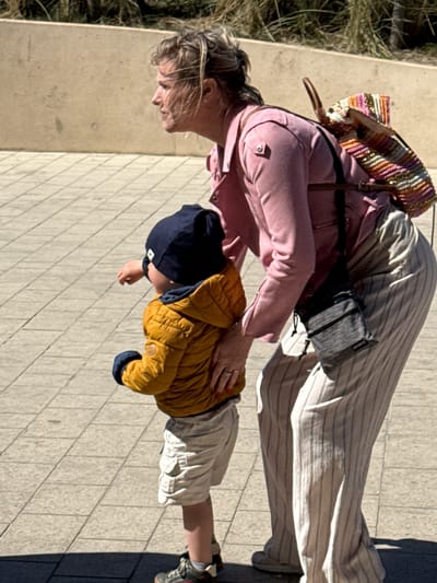 An outdoor, eye-level shot in Middelkerke, Belgium, shows a woman and a young child on a sunny day. The image is oriented vertically, but depicts subjects on a horizontal paved path.
The woman, with light-colored hair, is positioned on the left, bending forward and looking down. She wears a pink long-sleeved top or jacket, striped wide-leg pants (light beige or off-white with thin vertical stripes), and light-colored sneakers. A woven, multi-colored shoulder bag (pink, orange, gold, purple tones) is worn over her right shoulder, and a black fanny pack or crossbody bag, with the visible text "SLACKTIDE", is slung across her chest. A ring is visible on a finger of her left hand, which rests gently on the child's back.
To the woman's right is a young child, also bending slightly and looking downwards. The child wears a dark blue beanie with a small white square logo, a mustard yellow puffer jacket, light-colored shorts, and grey and orange sneakers.
They appear to be paused on a light-colored paved surface, possibly a sidewalk or promenade, composed of rectangular blocks. To the far left, beyond the paved area, is dry, light-colored vegetation, suggesting a natural border or landscaping. The strong shadows cast by the subjects indicate bright sunlight, suggesting midday or early afternoon. The interaction between the woman and child suggests a moment of care or guidance.