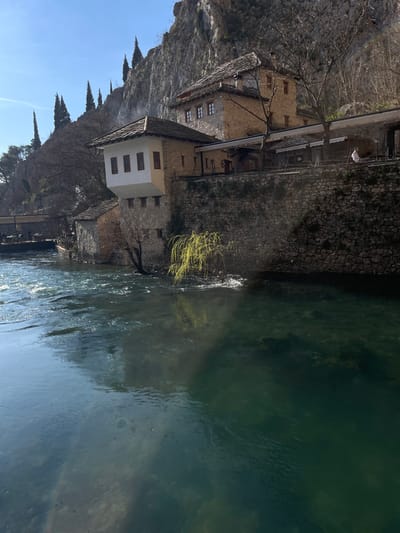 The image depicts an outdoor scene in Dračevice, Bosnia and Herzegovina, featuring a historic building complex nestled against a towering limestone cliff beside a swift-flowing, greenish-blue river. The primary architectural elements consist of stone and white-plastered buildings with traditional multi-gabled, stone-tiled roofs. A prominent lower building features a white, cantilevered upper section with three brown-framed windows, its stone foundation extending into the river. Above and to its right, a larger multi-story stone building with multiple windows stands against the cliff face. A stone-walled terrace or platform connects these structures, covered by dark awnings that shade an outdoor seating area equipped with tables and chairs. One individual in light-colored attire is seated at a table on this terrace, facing the river.
The environment includes the clear, flowing river occupying the foreground, displaying visible currents and surface reflections under direct sunlight. A weeping willow-like tree with bright green, newly unfurled leaves extends over the water near the lower building. The steep, grey-brown cliff dominates the background, topped with several tall, slender evergreen trees and interspersed with bare deciduous trees. The sky is clear and bright blue, indicating fair weather. A wooden bridge or walkway is partially visible on the far left, upstream.