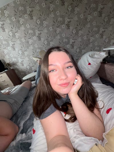The image shows a young woman lying on a bed in a room with floral wallpaper. She is looking at the camera with a slight smile, resting her head on her hand. She is wearing a grey t-shirt and grey shorts. The bed has a striped duvet with small red hearts. A straw hat is visible on one of her feet. A bedside table with drawers is in the background, along with a television. The room appears to be a bedroom in a residential setting. The location is identified as Bashkultaevo, Russia.
