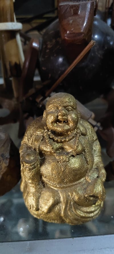 A golden-colored figurine of a seated, corpulent figure, consistent with Budai iconography, occupies the center foreground. The figure displays a wide, smiling mouth with visible teeth, closed eyes, and a prominent belly. A textured, metallic gold finish covers the figurine, showing surface irregularities. A beaded necklace is depicted on its chest. The figure's left hand holds a small, spherical object. It rests on a translucent surface, likely glass, which exhibits particulate dust. A grey border is visible beneath this surface. The background is blurred and dark, featuring various indistinct objects: a vertical, light-colored, possibly wooden element on the upper left, a dark, rounded vessel on the upper right, and a slender, reddish-brown stick extending diagonally across the upper right quadrant. The scene depicts an indoor display in Cikanyere, Indonesia.