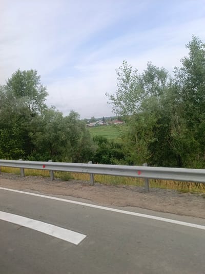 The image depicts a roadside view in Akulovo, Russia. A grey asphalt road with a white line marking is visible in the foreground, extending from the bottom left to the top right. A metal guardrail runs along the edge of the road, separating it from a grassy verge. Beyond the guardrail, a lush green landscape unfolds, with dense trees and vegetation. In the distance, a small village with houses and fields can be seen. The sky is overcast with light clouds. The scene is static and appears to be taken from a moving vehicle. No people or animals are present in the image. The lighting suggests it is daytime, likely morning or afternoon, with diffused light due to the cloudy conditions.