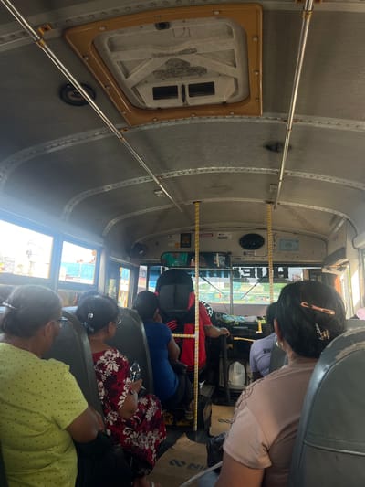 The image shows the interior of a crowded bus in Managua, Nicaragua, looking towards the front. Several passengers are seated, facing forward, with their backs to the camera. To the left, a woman in a lime green polka dot shirt is visible. Next to her, a woman in a red and white floral dress holds a bottle of water. In front of them, a man in a red shirt is operating the bus. The ceiling of the bus is gray and metallic, with visible rivets and overhead handrails. A yellow and white striped pole stands near the driver. The windows show a blurry view of buildings and street activity.