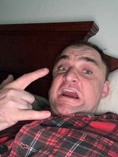 A close-up, overhead, first-person perspective shot shows a Caucasian man lying on a bed. The man is wearing a red and black plaid shirt and has a ring on his left hand. His eyes are open and he is making a distorted facial expression. His mouth is open and his tongue is visible. His left hand is raised and making a rock-and-roll gesture. The bed has a wooden headboard. The background is a plain wall. The image is likely taken in a bedroom in Lesnoi Gorodok, Russia, though this cannot be definitively confirmed from the visual information alone.