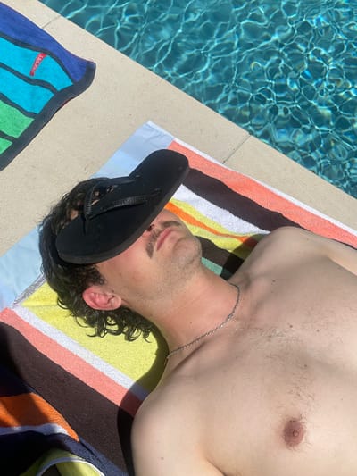 An individual is depicted lying supine on a multi-striped beach towel adjacent to a swimming pool in The Gap, Australia. A single black flip-flop rests horizontally across the individual's face, obscuring the eyes and forehead. The subject has dark, curly hair, a mustache, and is shirtless, wearing a silver-colored chain necklace. The towel beneath the individual features stripes in light blue, yellow, white, orange, and brown/dark orange. To the upper left, a portion of a second, folded striped towel displaying the word "ESPRIT" is visible. The bright blue water of the swimming pool, characterized by surface ripples and reflections, is situated immediately adjacent to the individual, separated by a light-colored concrete or tiled edge. Direct sunlight casts distinct shadows across the scene.