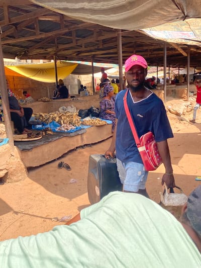 The image depicts a bustling outdoor market scene in Amaseri, Nigeria. A man in a blue t-shirt, light denim shorts, and a pink baseball cap is walking, carrying a black suitcase in his left hand and a red bag with a strap over his right shoulder. In the foreground, a person wearing a light green t-shirt is partially visible. The background shows stalls with various goods, including dried fish, and other people engaged in market activities under a covered structure with wooden beams and a corrugated roof. The ground is sandy and dusty, typical of an outdoor market environment in the region.