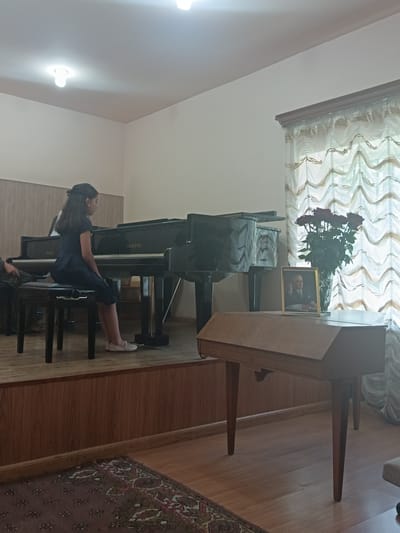 A young girl with dark hair pulled back is seated at a black grand piano, her feet resting on the pedal. She is wearing a dark blue dress and appears to be playing or about to play. The piano is on a raised wooden stage, with a textured wooden wall behind it. To the right of the piano, a vase of red roses sits on a wooden table, next to a framed portrait of a man in a suit. A sheer curtain with a wavy pattern hangs in the background. The scene is indoors, illuminated by ceiling lights. A patterned rug is visible on the floor in the foreground. The setting suggests a performance or practice session, possibly at a cultural center or home. The overall mood is calm and focused. Visible text on the piano reads "Estonia". The location is Nor Hachin, Armenia.