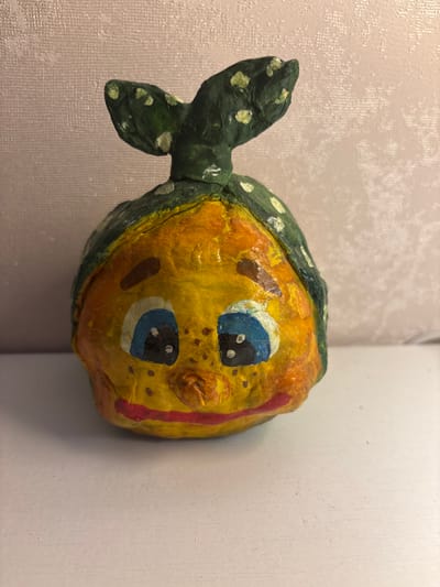 A handmade clay figurine of a cheerful character, resembling a round, yellow fruit with a green patterned headscarf, sits on a white surface. The character has large blue eyes with black pupils and white dots, a small brown nose, and a red mouth. It is adorned with brown freckles and two brown eyebrows. The green headscarf has white polka dots and two pointed ends that stick up like ears. The background is a textured, pale pink wall. The figurine appears to be in a domestic indoor setting. The city of Imeni Morozova, Russia is not depicted or suggested by the image.