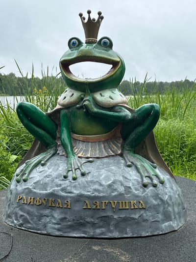 This is an upright, medium shot of a colorful statue of a frog wearing a crown, sitting on a rock. The frog is green with blue eyes and a wide, open mouth. It is wearing a beige cape and a white frilled skirt. The rock the frog is sitting on is grey and textured. There are gold-colored Cyrillic letters on the rock. To the left of the frog, there is a body of water with trees in the distance. The sky is overcast. The overall scene appears to be outdoors, possibly in a park or by a lake, given the presence of water, reeds, and grass. The setting is Местечко Раифа, Russia. The visible text on the rock reads "РАИФСКАЯ ЛЯГУШКА" which translates to "Raifa Frog".