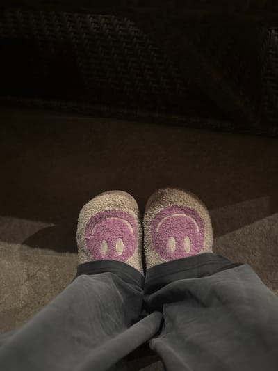 The image depicts a first-person, downward view of a person's lower extremities within an indoor environment in McAllen, United States of America. A pair of light-beige, plush slippers, each featuring a large pink embroidered smiley face with oval eyes and an upward-curving mouth, are worn on the feet. Dark grey casual trousers cover the lower legs and ankles, partially obscuring the top of the slippers. The subject stands or sits on a light brown tiled floor with visible grout lines. In the upper background, a dark, woven object, consistent with wicker furniture, casts a shadow across the floor. Ambient lighting illuminates the scene from an unseen source, creating soft shadows.