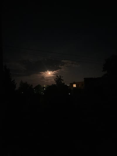The image captures a nighttime scene in the city of Stari Sanzhary, Ukraine. The sky is dark with scattered clouds partially obscuring a bright, orange-hued moon. In the mid-ground, silhouetted trees and foliage create a dark, foreground element. A building with a single illuminated window is visible on the right side of the frame. Overhead, power lines are visible against the night sky. The overall impression is one of a quiet, possibly rural, residential area at night.