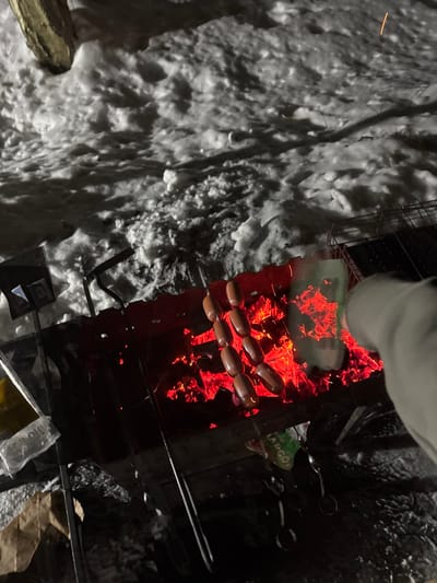 A bird's-eye view captures sausages grilling over an intense fire, the coals glowing a vibrant red. Snow surrounds the grill, creating a stark contrast between the cold exterior and the heated cooking area. Utensils and a package sit at the bottom. A partial view of a person's arm/sleeve.
