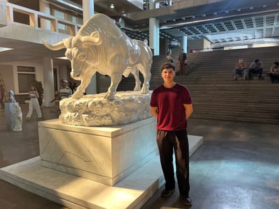 A young man stands in the foreground of a wide shot, posing for a photo in an indoor space. He is wearing a maroon t-shirt and black pants, with dark hair and a smile. To his left, a large, white sculpture of a bull is displayed on a multi-tiered marble base. The bull sculpture is detailed and appears to be made of plaster or a similar material. In the background, a grand staircase ascends, with several people seated on the steps and walking on the landings. Other sculptures and architectural elements are visible, including a statue of a woman to the far left. The setting appears to be a museum or public building, with bright overhead lighting. The time of day is not specified, but the lighting suggests it is indoors. The overall mood is casual and observant, with people interacting with the art and the space. The location is Greater Noida, India.