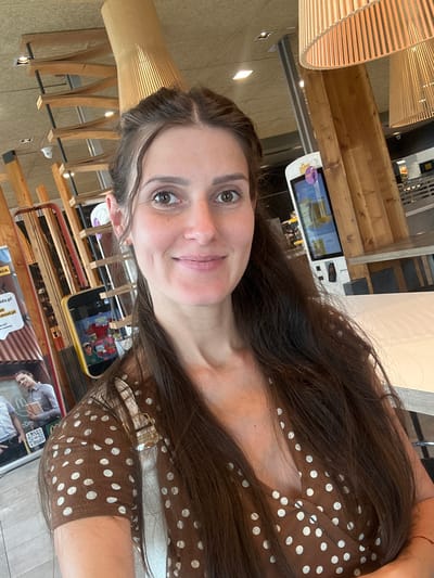This vertically oriented image features a young adult woman taking a selfie inside a modern fast-food restaurant or cafe in Ustroń, Poland. She has long, dark brown hair parted on the side and a pleasant, slightly smiling expression with brown eyes. She is wearing a brown top adorned with white polka dots and a thin, red string-like bracelet on her right wrist.
The indoor setting is characterized by extensive use of light-colored wooden elements, including large, circular slatted light fixtures and structural beams. In the background, multiple digital self-service kiosks are visible, displaying various advertisements. One screen shows a beverage, and another depicts content for children. Below the kiosks, promotional posters are displayed. Visible text on these posters includes a partial McDonald's logo ("Mcd") and Polish words like "Kids!". The bright, artificial lighting suggests a well-lit indoor environment. The woman appears calm and content.