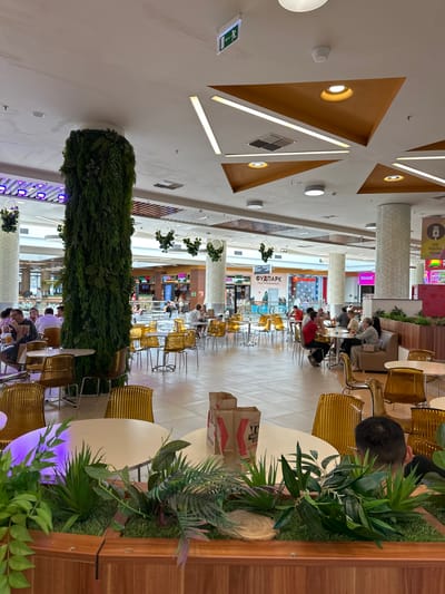 The image depicts a spacious, brightly lit food court area in a shopping center. A large, pillar-like structure covered in dense green foliage is a prominent central feature. Several tables with modern, translucent yellow chairs are arranged throughout the space, some occupied by diners. In the background, various food stalls with signage are visible, indicating different dining options. The ceiling is decorated with a geometric design of orange triangular panels and integrated lighting. An illuminated exit sign is visible on the upper left portion of the ceiling. Artificial plants and wooden planters are used as decorative elements within the food court. The overall atmosphere suggests a contemporary and bustling commercial environment. The location is identified as Kozulino, Russia.