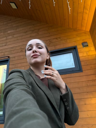 A young woman with light skin and dark hair pulled back is taking a selfie outdoors in front of a wooden building. She is wearing a dark green blazer over a brown shirt. Her left hand is resting on her chest, showing light blue nail polish and a ring on her left index finger. The building has horizontal wooden siding and a window with a dark frame. There are string lights hanging from the eaves of the building. The overall lighting suggests it is late afternoon or early evening. There is no indication of the specific location within Kamyennoye, Russia.