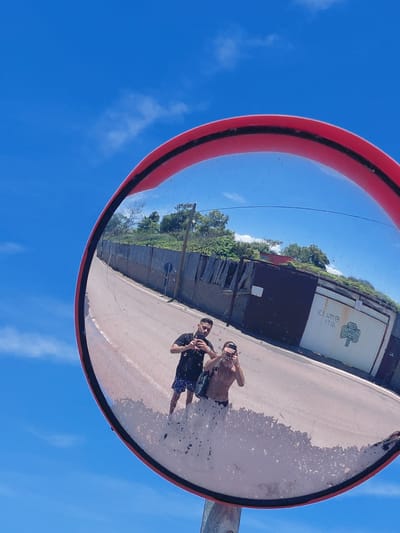 A wide-angle convex mirror reflects two individuals standing on a paved surface, likely a road, under a clear blue sky with scattered clouds. The mirror, with a red frame, is positioned outdoors, and its reflection captures a scene with a grey fence, a street, and buildings with overgrown vegetation. The first individual, on the left of the reflection, is wearing a black t-shirt and patterned shorts, holding a phone to take a photograph. The second individual, to the right, is shirtless and wearing dark shorts, also holding a phone. The reflection shows a building with a white facade bearing a green cloverleaf symbol and some indecipherable text. The paved surface in the reflection appears cracked and somewhat dusty. The specific location in the Municipal Unit of Aegio, Greece, cannot be determined from the visual information provided.