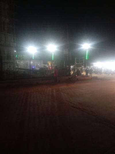 The image depicts an outdoor scene at night, illuminated by bright streetlights. The foreground is a dirt or gravel road, reflecting the light from above. In the midground, there is a dark metal gate with vertical bars. Beyond the gate, several people are visible, along with some parked motorcycles and what appear to be food stalls or temporary structures. The overall impression is of a bustling night market or gathering.
There are multiple bright lights, appearing to be street lamps or temporary lighting, casting a strong glow and creating starburst effects in the image. Two prominent lights are positioned on tall poles, one to the left and another slightly more to the right of the center. Smaller green lights are also visible, possibly decorative or part of the lighting setup. The sky is dark, indicating it is nighttime.
One person in the midground, to the right of the gate, is wearing a reddish-orange garment. Other figures are silhouettes against the bright lights, making their details indistinct. The atmosphere appears lively, suggested by the presence of many people and the activity implied by the stalls and parked vehicles. There is no visible text in the image. The setting suggests a public area, possibly a street or open space in a town or city, consistent with the provided location context of Tundun Wada South, Nigeria.