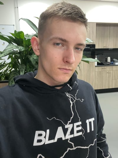 The image is a selfie taken indoors, likely in an office environment. A young man with short brown hair and blue eyes is looking at the camera. He is wearing a black hoodie with a white lightning bolt design and the text "BLAZE IT" printed on the front. To his left is a large green plant with variegated leaves. In the background, there are light-colored cabinets, a countertop, a black appliance that appears to be a toaster oven, and a silver thermos. The overall setting suggests a modern office break room or kitchen area. The specific location is not discernible from the image alone; it is not possible to determine if this is in Oldenzaal, Netherlands.