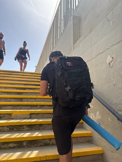The image depicts a bright, sunny day in Vilassar de Mar, Spain, with a group of people ascending a concrete staircase. The stairs are marked with yellow stripes on the treads. In the foreground, a person with a black baseball cap and a large black tactical backpack is walking up the stairs. They are wearing black shorts and a black t-shirt. The person is also carrying a long, blue cylindrical object, possibly a case or a rolled-up mat, in their right hand.

Behind this individual, two other people are also ascending the stairs. The person closer to the foreground is a woman wearing a black strapless top and dark shorts. She is wearing sunglasses and has her hair in a bun. The person further up the stairs is a man, partially visible, wearing dark shorts and appearing to be shirtless. The background features a beige concrete wall with a metal railing running alongside the stairs and a glimpse of a fence structure above the wall. The sky is clear and blue with a faint contrail from an aircraft. The lighting suggests it is midday.