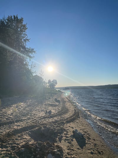 The image depicts a sunny day at a beach in Solnechnaya Polyana, Russia. The scene is oriented vertically, with the sky and sun on the left and the water and sand on the right. The bright sun is positioned in the upper left quadrant, casting a lens flare that extends diagonally across the image. The sky is a clear, vibrant blue, fading to a lighter shade near the horizon.
The water, a deep blue, meets the shore with small waves and sparkling highlights from the sun. The beach is composed of dark sand, with some scattered rocks and imprints, possibly from footprints. Lush green trees and foliage are visible along the edge of the beach, framing the scene. The overall atmosphere is serene and peaceful, suggesting a pleasant day outdoors. The time of day appears to be morning or late afternoon due to the sun's angle and the quality of light. No people or text are visible in the image.