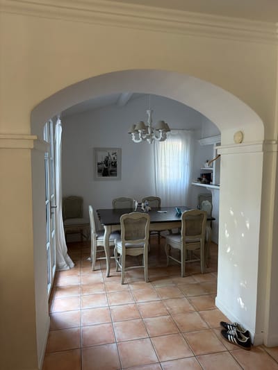 The image shows a dining room interior in a house, possibly located in Grimaud, France, given the architecture and style. The dining room is seen through an arched doorway with decorative molding. The room contains a rectangular dining table with at least six chairs arranged around it. The chairs have a light-colored, possibly wicker or cane back design and light-colored fabric seats. A chandelier hangs above the table, casting light onto the room. A framed picture hangs on the wall opposite the arched doorway. A window with white curtains is visible in the background, providing natural light. The floor is tiled with large, square terra cotta tiles. A pair of black and white sneakers are located near the doorway on the tiled floor. The architectural style suggests a Mediterranean or European design.