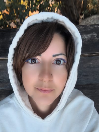 A medium close-up, head-and-shoulders portrait depicts an adult female positioned centrally, looking directly into the camera. The subject has dark brown, straight hair with bangs, partially visible beneath the hood. Her brown eyes are wide open, featuring a distinct application of blue mascara on the lower lashes. A gold-toned, C-shaped septum ring is visible in her nose, and a small dark mark is present on the left side of her nose. Her lips are closed with a slight upward curve. She is wearing a light-colored, likely off-white or light grey, hooded sweatshirt with the hood pulled over her head. The immediate background behind the subject consists of dark, weathered wooden planks arranged horizontally. The upper background features heavily blurred green and yellow foliage, indicative of an outdoor setting under natural light. The scene is located in the city of Shaalvim, Israel.