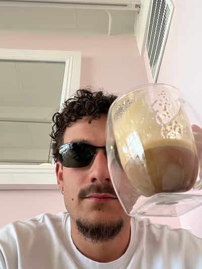 A man in sunglasses and a white t-shirt holds a glass of liquid in front of his face in an indoor setting with pink walls. He has curly hair and a beard. In the background, there is a vent and a mirror or door with a white frame. The location is likely a bathroom, as indicated by the mirror and vent. The exact location in Palm Desert, United States of America is unknown.