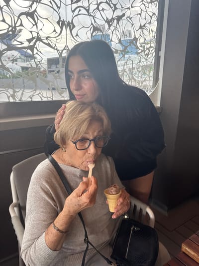 The image displays two females indoors at a location in the Berry Islands, Bahamas. An elderly woman with short, light brown hair, wearing black-framed eyeglasses, a light grey long-sleeved top, and a bracelet on her left wrist, is seated in a grey chair. She is actively eating chocolate-colored ice cream from a small cone with a wooden spoon. A black cross-body bag hangs from her shoulder. Standing immediately behind her is a younger woman with long, dark hair, dressed in a dark top. The younger woman's right hand rests gently on the elderly woman's head, and she gazes towards the left side of the frame. Behind them is a large window featuring an intricate, abstract grey and white wavy pattern. Through this window, multiple large white and blue cruise ships or yachts are visible, along with distant structures under an overcast sky, indicating a port environment. A dark wooden table edge is partially visible in the lower right foreground.
