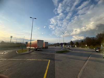 The image depicts a highway service area or truck parking facility located within the city of Radovljica, Slovenia, during late afternoon or early evening. The scene is illuminated by low-angle sunlight from the left, casting long shadows across the asphalt pavement.

In the foreground, dark asphalt with a yellow painted line is visible. The mid-ground features a road system with a prominent circular, grass-covered island. A green directional sign on this island points left, indicating "Jesenice." Two semi-trailer trucks are visible near this island; the lead truck has a red/orange trailer with a white roof, and the second truck's trailer is primarily white. Adjacent to the trucks are tall streetlights and various road signs, including a blue 'P' for parking and a triangular 'Give Way' sign (inverted triangle with red border).

To the right, an asphalt road curves, leading to a parking area where several passenger cars are present, with one dark-colored car partially visible on the far right edge of the frame. Additional streetlights line this road and parking zone.

The background consists of a horizon line of deciduous trees, mostly bare, beyond which a range of snow-capped mountains rises. The sky is partly cloudy, transitioning from a warm, bright blue on the left to a more overcast, deeper blue with white cumulus clouds on the right. No individuals are distinctly visible within the frame.