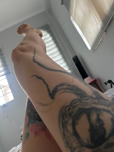 The image displays a close-up view of a person's leg, adorned with several tattoos. The leg is positioned in a relaxed pose, with the foot extended upwards. The background suggests an indoor setting, possibly a bedroom, with a window featuring blinds and natural light streaming in. There are no discernible elements in the image that can be used to determine the location of the city of Buenos Aires, Peru, as this is not a recognized geographical location. The scene is devoid of any contextual information about the environment or the subjects involved.