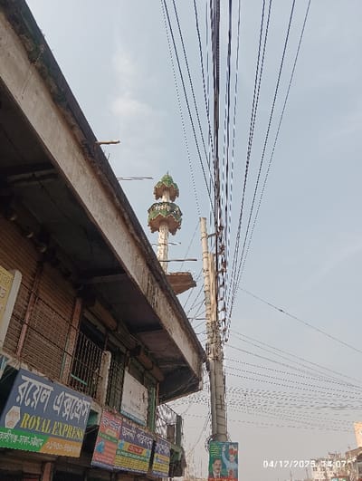 The image depicts an upward-angled view of a street scene in Kaliganj, Bangladesh, under a bright, hazy sky. On the left, the weathered concrete facade of a commercial building is visible. This structure features multiple storefronts with rolled-down metal shutters and security grilles. Prominently displayed is a blue sign in Bengali and English reading "ROYAL EXPRESS," with additional Bengali text indicating routes like "Kaliganj-Dhaka-Chittagong" and a phone number. A second partially visible sign reads "জননী পরিবহন" (Jononi Poribohon). Behind the building's facade and slightly to the right, a white minaret with a distinct green and white tiered, decorative top section rises. To the right of the minaret and extending towards the right edge of the frame, a robust concrete utility pole stands, laden with a dense array of black electrical and communication wires fanning out across the sky. A green and white political poster, depicting two male figures, is affixed to the lower section of this utility pole. A digital timestamp "04/12/2023 14:07" is present in the lower right corner. No specific individuals or direct actions are discernible within the frame.