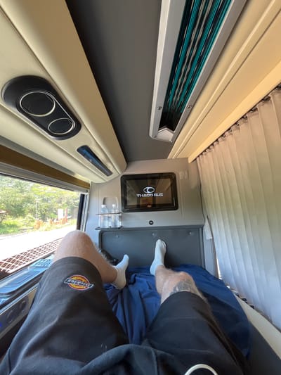The image shows a first-person view of a person lying down on a bed inside a bus cabin. The person is wearing black shorts with a Dickies logo and white socks. The cabin is equipped with a television screen displaying the "THACO BUS" logo, air vents, and a window with a view of trees and railroad tracks. There are also two bottles of water on a shelf. The overall impression is of a comfortable and private resting space within a bus, likely on a journey from or to Phú Hòa, Vietnam.