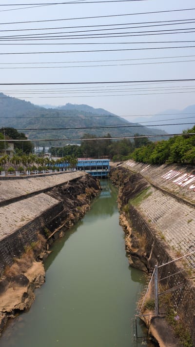 This aerial-perspective photograph shows the Dinh Dinh dam and the surrounding landscape in Lâm Sơn, Vietnam. The dam is constructed of grey stone blocks and concrete. A blue building sits near the water's edge. The water is a murky greenish-brown. In the distance, a mountain range is visible under a hazy sky. Power lines crisscross the left side of the image. The camera appears to be positioned above the water level, offering a view down into the dam and the surrounding terrain.