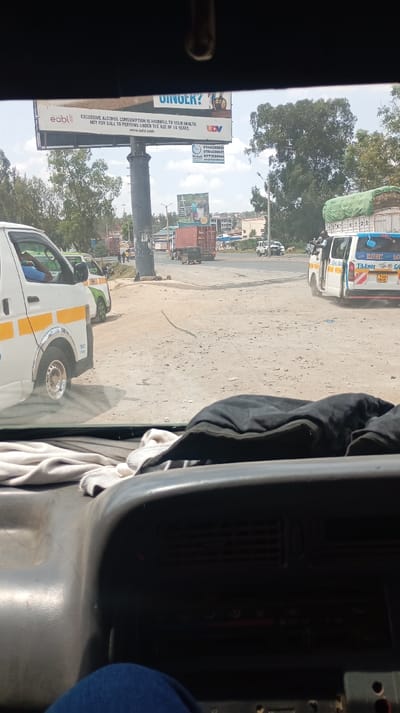The image shows a view from inside a vehicle, looking out onto a dusty intersection with a prominent billboard in the background. The billboard advertises an alcoholic beverage and warns against underage drinking and excessive alcohol consumption. Several white vans with yellow stripes are visible, and a semi-truck is further down the road. The dashboard of the vehicle dominates the lower portion of the frame, with some clothing items placed on it.