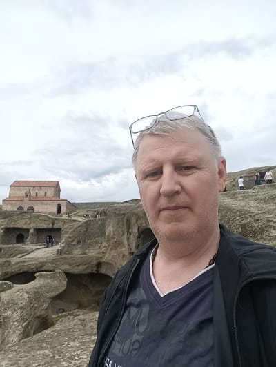 The image is a selfie taken by a man with graying hair and a beard, wearing glasses on top of his head. He is looking directly at the camera. He is wearing a black jacket over a dark blue V-neck t-shirt with a white layer underneath. A beaded necklace is visible around his neck.
The background depicts the ancient cave city of Uplistsikhe, Georgia. In the mid-ground, a stone church with a red roof stands prominently on a hillside. The foreground and mid-ground are characterized by the rocky, carved landscape of the cave city, with various openings and structures carved into the rock. Several other people are visible in the distance, some standing near the church and others exploring the cave dwellings. The sky above is overcast.