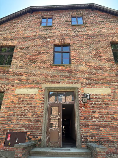 The image shows a close-up of a weathered brick building, likely an old industrial or institutional structure. The building has a dark, sloped roofline on the left, with a wooden fascia. Several windows are visible; some are small and square with multiple panes, reflecting the sky, while others are larger and rectangular, featuring darker frames and panes that reflect the interior and exterior of the building. One prominent doorway is open, revealing a dark interior with some barred elements.
The brickwork is a mix of reddish-brown and darker shades, showing signs of age and wear. There are also sections of lighter-colored plaster or concrete integrated into the wall. A small, dark brown electrical box with warning symbols and labels is mounted on the brickwork near the open doorway. Above the doorway, a sign with a black background and white numbers "3" and "13" is attached to the wall, possibly indicating a room or section number. The overall impression is one of a historic, possibly utilitarian building with a sense of history. The lighting suggests it is daytime, with clear skies. The context of Oświęcim, Poland, suggests this could be a building within the Auschwitz-Birkenau Memorial and Museum complex, given the architectural style and the somber atmosphere often associated with such locations.