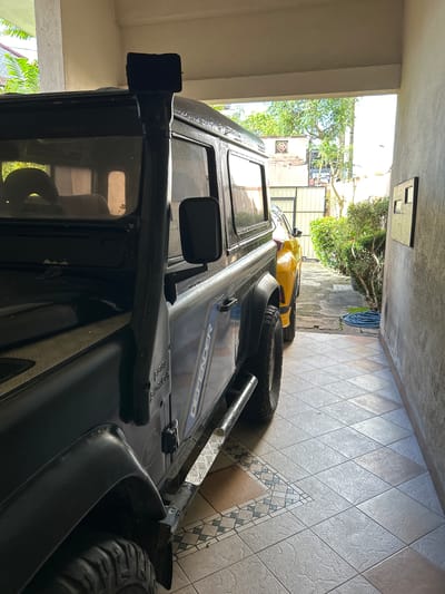 The image depicts a narrow, covered outdoor walkway or garage space in Dehiwala, Sri Lanka. Two vehicles are parked within this space. In the foreground and to the left, a dark-colored, off-road vehicle, likely a Land Rover Defender, is prominently featured. Its snorkel attachment on the roof and the "DEFENDER" decal on the side are visible. The vehicle has a rugged, utilitarian appearance with large tires and a metal running board. To the right of the Defender and further into the background, a bright yellow compact SUV or hatchback is parked. The walkway has a tiled floor with a decorative inlay pattern. Lush green vegetation is visible beyond the open end of the walkway, along with parts of buildings and trees, suggesting a residential or commercial area. The lighting indicates it is daytime, with sunlight illuminating the scene. There are no people visible in the image. A blue garden hose is coiled on the ground to the right. A black electrical box or panel is mounted on the wall to the right.