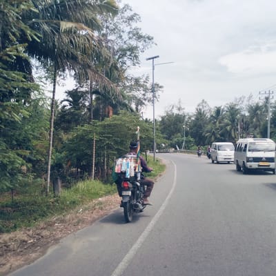 A street vendor on a motorcycle, viewed from behind, drives along a paved road lined with lush tropical vegetation. The vendor's motorcycle is heavily laden with goods for sale, including numerous colorful plastic bottles or pouches, likely drinks or snacks, hanging from a rack on the back. A multi-colored umbrella is also attached vertically to the motorcycle. The vendor wears a dark shirt and pants with a black cap.

The road curves gently to the right, with a white solid line marking the shoulder. On the left side of the road, dense foliage consisting of various palm trees, other trees, and bushes forms a green wall. On the right side, the road verge is grassy, giving way to more trees in the distance.

Further down the road, two white vans or minibuses are visible, traveling in the opposite direction. The license plate on the closer van reads "BL 1293 CZ." Other smaller motorcycles can be seen in the distance. A street light with a solar panel is positioned on the left side of the road, and utility poles with power lines are visible on the right. The sky is bright and overcast, indicating daytime. The scene depicts a typical daily activity in a semi-rural or rural area, consistent with the tropical climate and infrastructure of Cotbada, Indonesia.
