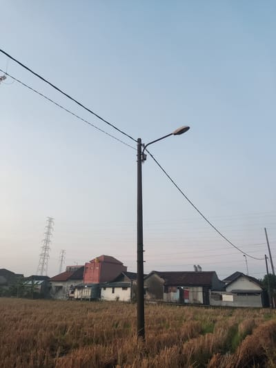 The image depicts a rural scene in Sukorejo, Indonesia, at dawn or dusk. A tall, dark lamppost stands in the foreground, its light fixture angled towards the right. Multiple power lines crisscross the sky above, some attached to the lamppost and others extending towards distant electrical towers visible on the left side of the frame. In the middle ground, a row of houses with various roof styles and materials is situated behind a field of dry, golden straw, likely remnants of a harvested rice paddy. The houses appear to be simple, residential structures with some showing signs of wear and tear. The sky is a pale, clear blue, indicating calm weather conditions. No people are visible in the image.