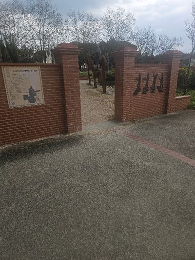 This image depicts an outdoor scene in Avignonet-Lauragais, France, specifically an entrance to a public area or memorial. In the foreground, a dark gray, pebbled pavement occupies the lower half of the frame, with a thin diagonal strip of reddish-brown bricks bordering a section of the pavement on the right.
The midground features an open gateway formed by two sections of a low brick wall, each capped with a brick pillar. The wall on the left displays a light-colored rectangular sign with text and a dark silhouette of a person playing a drum kit. The wall section to the right of the entrance features four dark silhouettes of individuals playing brass instruments, depicted in a marching or performing posture.
Through the brick gateway, a light-colored gravel path is visible, leading into an area with a series of vertical, weathered, dark-colored structures or sculptures arranged in a line along the path. Beyond these structures, the background consists of numerous deciduous trees with mostly bare branches, intermingled with some evergreen foliage. The sky above is overcast with visible cloud cover. No individuals are present in the immediate vicinity.