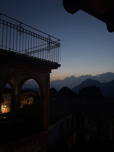An evening shot captures a view from a balcony in Orino, Italy. The sky is a deep twilight blue, transitioning to lighter shades near the horizon where soft clouds are silhouetted against the fading light. In the foreground, a dark, ornate metal railing of a balcony runs along the top of the frame, supported by a brick structure with arched openings. Through one of these arches, a warm, interior light illuminates a small section of the wall. Below, the roofs of several buildings are visible, cloaked in shadow. Beyond the immediate structures, dark trees are scattered, and in the distance, a range of mountains fades into the dusky atmosphere. Faint lights from a distant settlement can be seen on the slopes.
