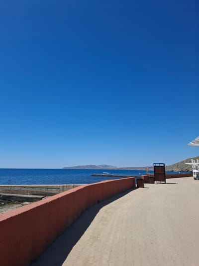 This image depicts a seaside promenade on a bright, clear day. The scene is dominated by a vast expanse of deep blue sky. To the right, a paved walkway with a textured, light-colored surface stretches into the distance, bordered by a low, reddish-orange wall. Beyond the wall, the calm, blue sea meets a distant, hazy shoreline. In the upper right portion of the image, there are structures that appear to be part of a waterfront area, possibly including seating or railing elements. The orientation of the image is sideways. The clear blue sky and bright light suggest it is daytime, likely mid-morning or afternoon, with fair weather. The setting appears to be a coastal town or resort area. No people or specific activities are evident, giving the scene a serene and tranquil feel. There is no visible text in the image.